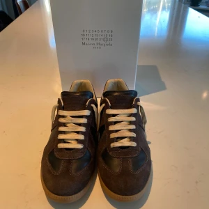 Maison Margiela Replica sneakers brun - Säljer ett par Maison Margiela Replica sneakers i mörkbrunt mocka och läder med vita skosnören och beige sula. Klassisk low-top siluett med retrovibbar och diskret branding på plösen. Perfekta för dig som gillar stilrena och tidlösa sneakers.