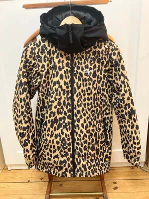 Leopardmönstrad skidjacka  från CLWR - Snygg skidjacka i nyskick från CLWR med leopardmönster i beige och svart. Jackan har svart huva, dragkedja framtill och två fickor med dragkedja. Perfekt för dig som vill sticka ut med en cool och trendig look. Snölås i midjan och ficka för liftkort! Nypris: 2.200kr