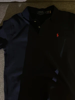 Marinblå piké från Polo Ralph Lauren - Klassisk marinblå pikétröja från Polo Ralph Lauren med röd broderad logga på bröstet. Modellen är Classic Fit och har korta ärmar, krage och knappar vid halsen. Perfekt för en stilren och avslappnad look. Helst oanvänd men ingen prislapp på!