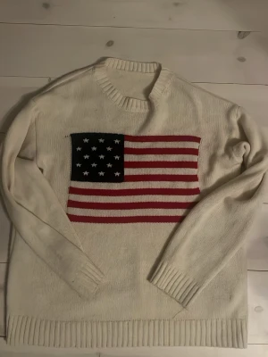 Vit stickad tröja med USA-flagga - Vit stickad tröja med rund hals och ribbade muddar. På bröstet finns en stor amerikansk flagga i rött, vitt och blått. Tröjan har en klassisk passform och är perfekt för dig som gillar statement-plagg.