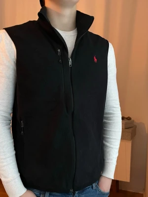 Svart Ralph Lauren fleeceväst, storlek L - Svart Ralph Lauren fleeceväst med röd logga och bra fickor.   100% polyester  Mycket bra skick, som ny  Storlek L men något mindre (passar M och han på bilden som är 184 väger 80) Längd: 68 cm