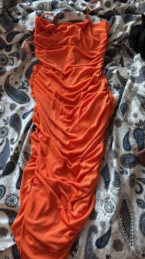 Orange långklänning med rynk - Säljer en snygg orange långklänning med tunna axelband och rynkade detaljer längs hela klänningen. Klänningen har en figurnära passform och är tillverkad i ett mjukt, stretchigt material som känns bekvämt mot huden. Perfekt för dig som vill sticka ut.