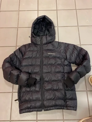 Svart dunjacka från Peak Performance - Svart dunjacka med huva från Peak Performance. Jackan har dragkedja framtill, två sidofickor med dragkedja och muddar vid ärmsluten. Perfekt för kalla vinterdagar och har en stilren, sportig look.