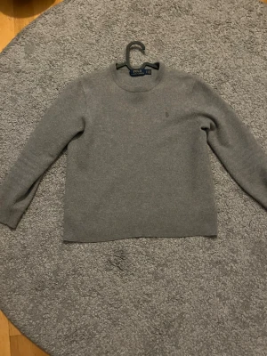 Grå stickad tröja i ullmix från Ralph lauren  - En stilren grå stickad tröja från Ralph lauren med rund hals och långa ärmar. Tröjan har ribbstickade muddar vid ärmslut och nederkant, vilket ger en snygg och enkel look. Perfekt att matcha med jeans eller kjol för en avslappnad stil.