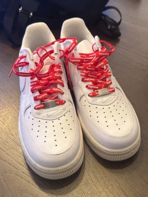 Nike Air Force 1 x Supreme vita sneakers - Säljer ett par vita Nike Air Force 1 sneakers i samarbete med Supreme. Skorna har röda skosnören med Supreme-logga och en liten Supreme-tag på sidan. Klassisk låg modell i läder med perforerad tå och Nike Air-detaljer på hälen.