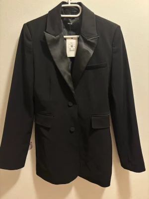 Svart kavaj med satinrevers från Bik Bok - Stilren svart kavaj från Bik Bok med glansiga satinrevers och två knappar framtill. Klassisk passform med fickdetaljer och bröstficka. Perfekt för att lyfta din outfit med en elegant touch. Materialet är slätt och ger en dressad känsla.