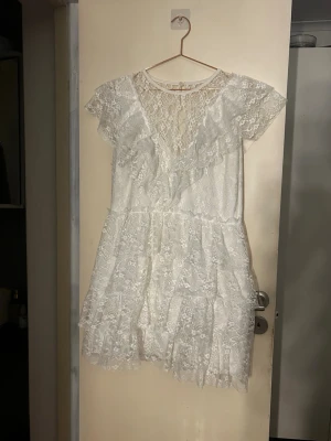Vit spetsklänning med volanger - Söt vit kortklänning i spets med volangdetaljer och v-ringad rygg. Klänningen har korta ärmar och ett romantiskt mönster. Perfekt för sommarens alla tillfällen och ger en fräsch och feminin look. Använd en gång. Perfekt till studenten. Står ej storlek men gissar på L