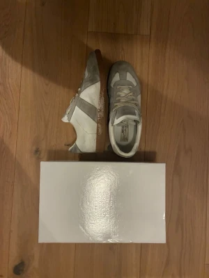 Vita och grå Maison Margiela sneakers - Maison Margiela sneakers i vitt och ljusgrått läder och mocka med klassisk gummisula. Snygg retrostil med lågt skaft och diskreta detaljer. Perfekta för dig som gillar minimalistisk design och streetwear-vibe.