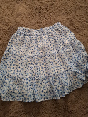 Vitblå blommig volangkjol - Söt kort kjol med blått blommönster på vit botten. Kjolen har flera volanger och resår i midjan för en bekväm passform. Perfekt för dig som gillar en lätt och luftig stil.