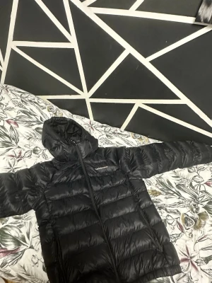 Svart dunjacka med huva från Peak Performance, Storlek M - Snygg svart dunjacka från Peak Performance med huva och dragkedja framtill. Jackan har en puffig design, två sidofickor med dragkedja och diskret logga på bröstet. Perfekt för kalla dagar och enkel att matcha med olika outfits.