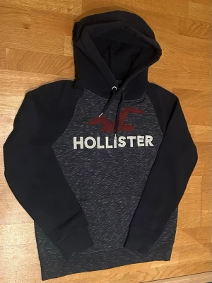 Mörkblå Hollister hoodie med logga - Snygg mörkblå hoodie från Hollister med stor vit logotyp och röd fågel framtill. Tröjan har huva med dragsko och känguruficka. Ärmar och nederkant har ribbade muddar. Perfekt för en avslappnad och trendig stil.