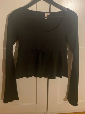 Svart långärmad topp med knyt - Svart långärmad topp från Nelly med rund halsringning och dekorativ knytning i midjan. Toppen har lätt utsvängda ärmar och vågig nederkant för en trendig look. Perfekt att styla med jeans eller kjol. Använt fåtal gånger och säljer pga att den inte kommer till användning längre❣️❣️