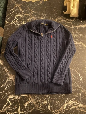 Ralph lauren quarter zip knit - Mörkblå kabelstickad tröja från Polo Ralph Lauren med halv dragkedja och röd broderad logga på bröstet. Tröjan har lång ärm, ribbstickade muddar och hög krage. Perfekt för lager-på-lager och klassisk stil. Det står L men det Är storlek S vet eh värför