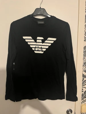 Svart långärmad topp Emporio Armani - Snygg svart långärmad topp från Emporio Armani med det ikoniska vita örnlogotypen och GA-tryck på bröstet. Klassisk rund halsringning och stilren design som passar till mycket. Tillverkad i mjukt material för skön känsla. Storlek xs (170) barnstorlek