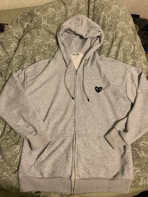 Grå hoodie från Comme des Garçons Play - Snygg ljusgrå hoodie med dragkedja från Comme des Garçons Play. Har huva med dragsko, två fickor fram och det ikoniska svarta hjärtat med ögon broderat på bröstet. Perfekt för en chill och stilren look. Storlek m