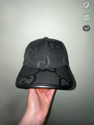 Svart keps från Gucci med GG-mönster - Snygg svart keps från Gucci med det klassiska GG-mönstret över hela kepsen. Kepsen har böjd skärm och detaljer i svart läder runt kanten. Perfekt accessoar för dig som vill ha en exklusiv och trendig look.