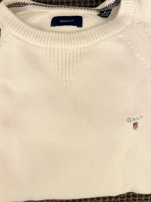 Vit stickad tröja från GANT - Klassisk vit stickad tröja från GANT med rund halsringning och diskret broderad logotyp på bröstet. Tröjan har ribbstickade muddar och detaljer vid halsen. Perfekt för en stilren och avslappnad look. Storlek 158/164cm