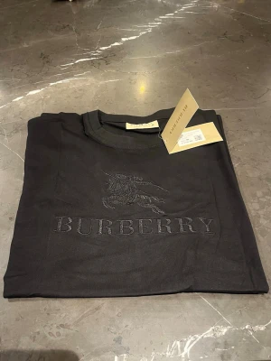 Svart Burberry t-shirt med broderi - Snygg svart t-shirt från Burberry med diskret broderad logga och motiv på bröstet. Klassisk rund halsringning och mjukt bomullsmaterial. Perfekt för dig som gillar stilrena designer och exklusiva märken.