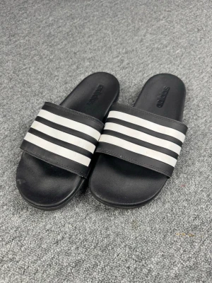 Svarta Adidas badtofflor med vita ränder - Klassiska svarta badtofflor från Adidas med tre vita ränder över foten. Tillverkade i syntetmaterial med räfflad sula för bra grepp. Perfekta för stranden eller gymmet och har en enkel slip-on design.