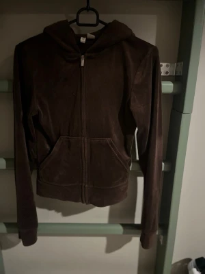 Mörkbrun velour hoodie med dragkedja - Mjuk mörkbrun hoodie i velour med dragkedja framtill och två stora fickor. Tröjan har huva och långa ärmar, perfekt för en chill och trendig stil.