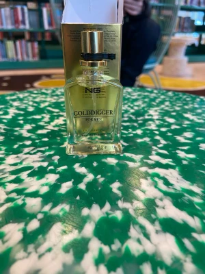 NG Golddigger For Men Eau de Toilette - Golddigger For Men från NG är en eau de toilette med maskulin doft. Flaskan är genomskinlig med guldfärgad vätska och har en elegant, fyrkantig form. Förpackningen är guldglänsande med svart text och bikakemönster, vilket ger en lyxig känsla.