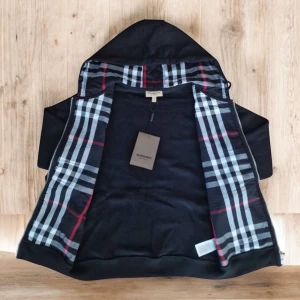 Svart Burberry hoodie med rutig huva - Svart hoodiejacka från Burberry med dragkedja, huva med klassiskt rutig mönster i svart, vitt och rött. Jackan har snörning i huvan, två fickor fram och ribbade muddar. Perfekt för dig som vill ha en stilren och ikonisk look.