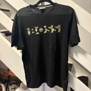 Svart BOSS t-shirt med tryck - Svart t-shirt från BOSS i storlek M med ett stort, guldigt BOSS-tryck på bröstet. Klassisk rund hals och korta ärmar. Materialet är mjuk bomull som känns skönt mot huden. Perfekt för dig som gillar stilrena logotyper och enkel design.
