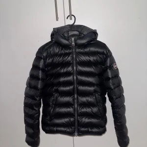 Svart dunjacka från Colmar med huva - Snygg svart dunjacka från Colmar med glansig finish och huva. Jackan har en klassisk pufferstil, dragkedja framtill och två fickor med dragkedja. Märkeslogga på ärmen ger en clean och sportig vibe. Perfekt för kalla dagar när du vill hålla stilen.