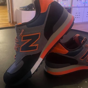 New Balance 576 Three Peaks sneakers - Snygga New Balance 576 Three Peaks sneakers med grå mesh, svart mocka och orange detaljer. Skorna har rund tå, platt sula och randiga orange/svarta snören. Insidan har ett unikt mönster med blå och orange accenter. Perfekt för dig som gillar streetstyle och färg.