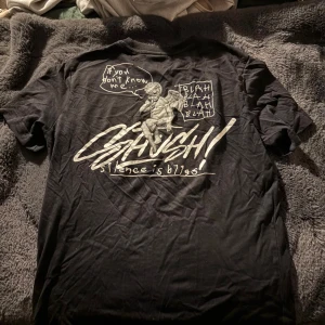 Svart t-shirt med tryck och text - Svart t-shirt med coolt vitt tryck på ryggen med texten 'SHUSH! silence is bliss' och en tecknad figur. Framsidan har en mindre version av samma tryck på bröstet. T-shirten har rund hals och korta ärmar, perfekt för en avslappnad streetstyle.