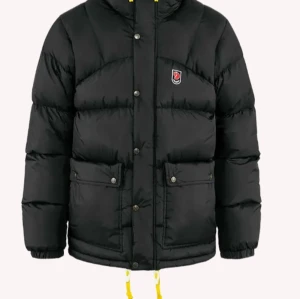Svart dunjacka från Fjällräven - Svart dunjacka från Fjällräven med klassisk pufferlook, gula snören och tryckknappar framtill. Jackan har två stora fickor med lock och en liten logotyp på bröstet. Perfekt för kalla dagar när du vill hålla stilen. Skriv för fler bilder!