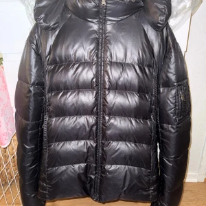  Hugo Boss - Säljer en svart dunjacka från Hugo Boss med glansig finish och lila foder. Jackan har huva, dragkedja framtill och quiltad design. Insidan är markerad med 'real down' och har en innerficka med dragkedja. Perfekt för kalla dagar och riktigt snygg . Kemtvätt utfördes 15/10. Fint skick 