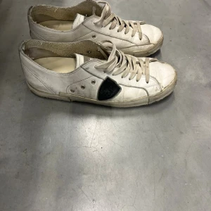 Vita sneakers med svart detalj - Säljer ett par vita Philippe Model sneakers med snörning och svart skölddetalj på sidan. Skorna har rund tå, platt sula och grått parti baktill. Tillverkade i skinn med slitna kanter för en cool, avslappnad look. Box följer med