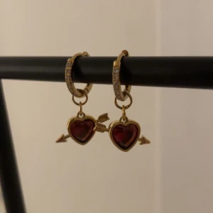 Guldörhängen med röda hjärtan och pil - Säljer ett par snygga guldörhängen med röda hjärtformade hängen och pil-detaljer. Ringen är dekorerad med små glittrande stenar som ger extra bling. Perfekt för dig som gillar romantiska och unika smycken med lite edge.