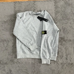 Grå sweatshirt från Stone Island - Snygg grå sweatshirt från Stone Island med klassisk rund hals och ribbade muddar. Tröjan har den ikoniska loggan på ärmen och är tillverkad i mjuk bomull. Perfekt för en clean och stilren look.