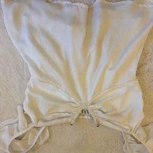 Vit omlott tubtopp från HWARE - Säljer en vit tubtopp från HWARE med snygg omlott-design och snörning framtill. Toppen är axelbandslös och har ett mjukt, lätt material som känns skönt mot huden. Perfekt för dig som gillar stilrena och trendiga plagg med coola detaljer.