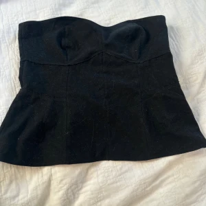 Svart korsettopp från Zara Woman - Säljer en svart axelbandslös korsettopp från Zara Woman. Toppen har en markerad byst, lätt utsvängd nederdel och dragkedja i sidan. Perfekt att styla med jeans eller kjol för en trendig look.