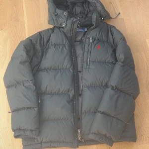 Svart dunjacka från Polo Ralph Lauren - Svart dunjacka från Polo Ralph Lauren M-L med röd broderad logga på bröstet. Jackan har huva, dragkedja framtill, snörning i nederkant och en bröstficka med dragkedja. Perfekt för kalla dagar och riktigt skön att bära. OBS på 3e bilden kan man se att ett av snörena är av 