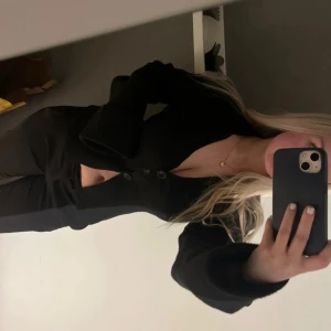 Svart kofta från Gina Tricot - Säljer en svart kofta från Gina Tricot med v-ringning och två knappar framtill. Modellen är  figursydd med långa ärmar och breda muddar med knappdetalj. Perfekt att styla med höga jeans eller kjol för en clean look.