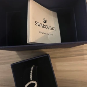 Swarovski armband med hjärta - Snyggt armband från Swarovski i silverfärg med glittrande stenar längs hela kedjan och ett öppet hjärta som hängsmycke. Justerbar kedja med ikonisk svanlogga på låset. Perfekt för dig som gillar eleganta detaljer och bling.
