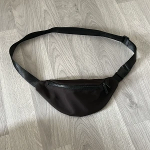 Svart midjeväska med dragkedja - En stilren svart midjeväska med justerbart band och smidig dragkedja framtill. Perfekt för att bära mobilen, nycklar och andra småsaker. Väskan har enkel design och är tillverkad i syntetmaterial med ett stort fack. Passar dig som vill ha händerna fria. Unisex.