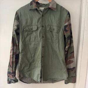 Ralph lauren skjorta M - Denna skjorta från Denim & Supply Ralph Lauren har en unik design med en kombination av enfärgad olivgrön framsida och kamouflagemönstrade ärmar och rygg. Skjortan har två bröstfickor och en klassisk krage. På ryggen finns en applikation med den amerikanska flaggan.  I väldigt gott skick!