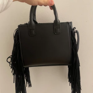 Svart handväska med fransar - Cool svart handväska med fransar på sidorna. Har slitage på handtagen därav billigt pris. Går säkert att fixa handtagen.