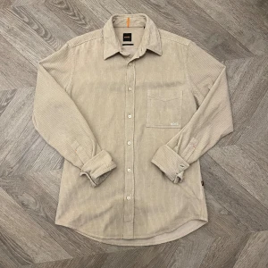 Beige manchesterskjorta från BOSS - Stilren beige skjorta från BOSS i mjuk manchester med diskreta ränder. Klassisk krage, bröstficka med broderad logga och knäppning framtill. Långärmad modell med avslappnad passform, perfekt för en clean och trendig look. Nypris 1399kr, använd fåtal gånger. 