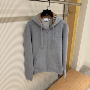Grå hoodie från Burberry med dragkedja - Snygg grå hoodie från Burberry med klassiskt rutigt foder i huvan. Tröjan har dragkedja framtill, två fickor och justerbar huva med snörning. Tillverkad i mjuk bomull och perfekt för chill dagar. Broderad Burberry-logga på bröstet.