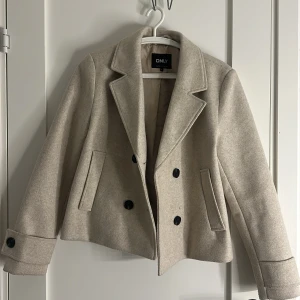 Beige kappa från ONLY - Snygg beige kappa från ONLY med klassisk krage, dubbelknäppning och svarta knappar. Jackan har två fickor fram och är lite croppad i modellen.