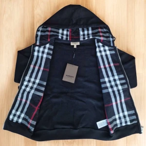 Svart hoodie från Burberry med dragkedja - Snygg svart hoodie från Burberry med klassiskt rutigt foder i huvan. Tröjan har dragkedja framtill, snörning vid huvan och två fickor. Tillverkad i mjuk bomull och har långa ärmar. Perfekt för dig som vill ha en stilren och exklusiv look.