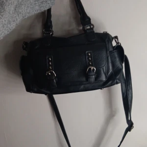 Svart axelväska med spännen - Säljer en svart axelväska i skinnimitation med både handtag och lång axelrem. Väskan har snygga metalldetaljer och spännen framtill samt dragkedja på baksidan. Perfekt storlek för att få plats med allt du behöver och passar till många olika outfits.