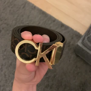 Brunt Calvin Klein bälte med guldspänne - Snyggt brunt bälte från Calvin Klein med klassiskt CK-mönster och ett stort, glansigt guldspänne. Bältet är gjort i skinn och har en stilren design som lyfter vilken outfit som helst. Perfekt för dig som gillar att sticka ut med detaljer.