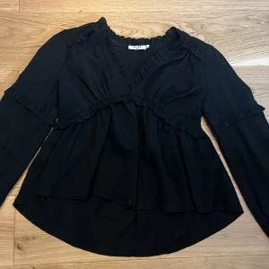 Svart volangblus från NA-KD - Svart blus från NA-KD i storlek 36 med volangdetaljer längs halsringning, ärmar och midja. Blusen har omlott-look framtill, långärmade puffärmar med resår vid ärmslut och är tillverkad i ett lätt, flowy tyg som ger en snygg siluett. Har använts 4 gånger.